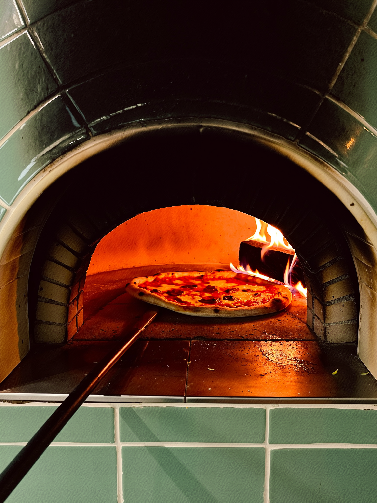 Een pizza gaat op een lange schep de hete houtoven in, vlammen likken langs de zijkant.