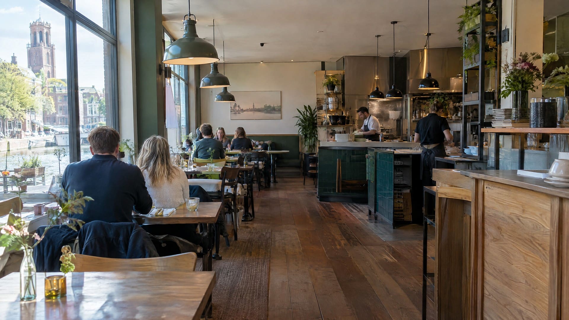 Eetzaal van een Italiaans restaurant met open keuken, donkergroene tegels, houten tafels en hoge ramen aan het water.